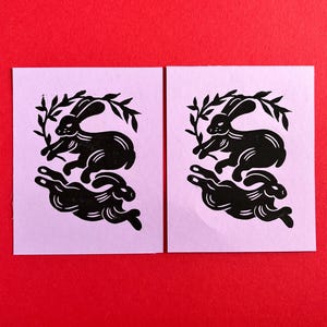 May include: Two black and white linocut prints of rabbits running in a circle with a leafy branch behind them. The prints are on a light purple background.