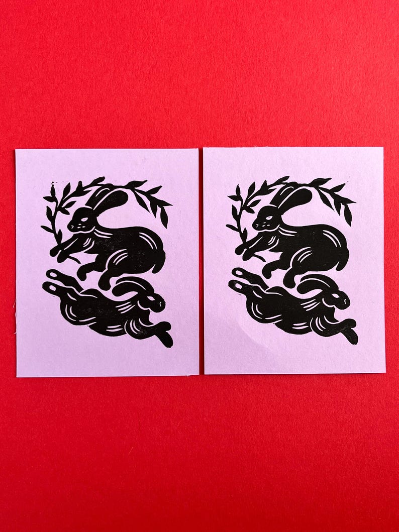 May include: Two black and white linocut prints of rabbits running in a circle with a leafy branch behind them. The prints are on a light purple background.
