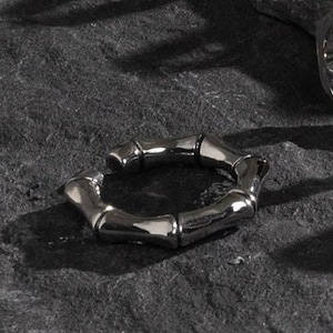 Puede incluir: Un anillo plateado con diseño de bambú. El anillo está hecho de múltiples segmentos conectados, creando una apariencia texturizada. Se encuentra sobre una superficie oscura y texturizada, con otro anillo visible en el fondo.