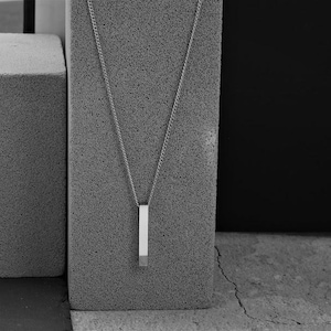 May include: A silver chain necklace with a rectangular pendant. The pendant is made of a silver bar with a smooth, polished finish. The necklace is displayed on a gray textured surface.