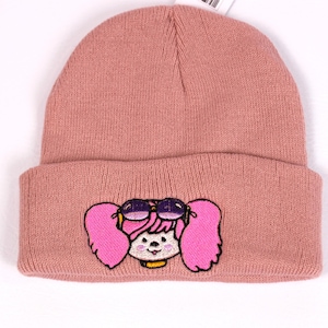 May include: A dusty rose beanie hat with a folded brim. The hat features an embroidered cartoon face with pink pigtails and sunglasses. The face has a smiling expression and a yellow collar.