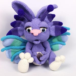 May include: A handmade crochet creature in shades of purple, blue, and green. The amigurumi has large ears, wings, and a pink nose. It is holding a small, dark-colored stone. The creature's feet are white.