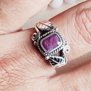 Pode incluir: Um anel de prata com uma gema roxa engastada em um bezel quadrado. O anel é decorado com detalhes em forma de folhas.