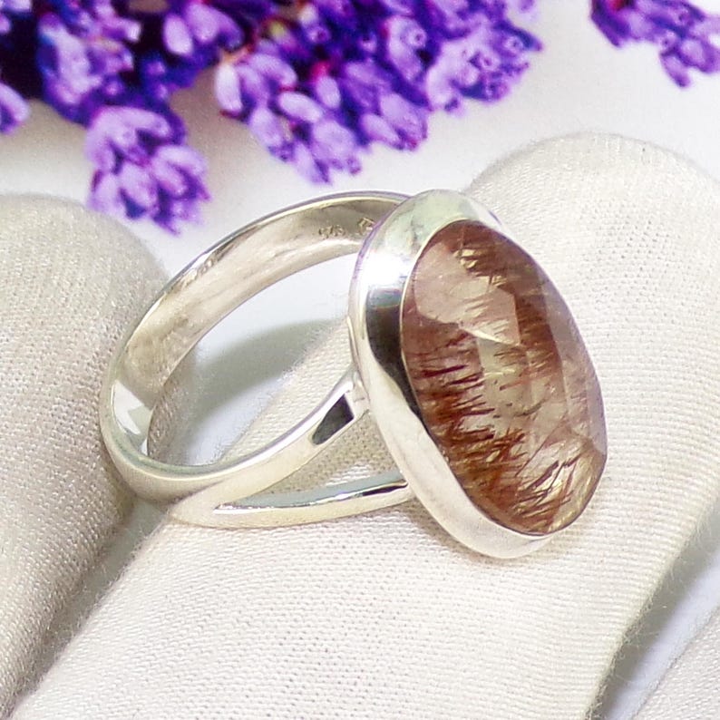 May include: A silver ring with a large, oval-shaped gemstone. The gemstone is a mix of brown and clear hues with internal lines. The ring is set on a white surface, with purple flowers in the background.
