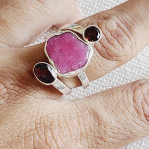 Peut inclure: Une bague en argent avec une grande pierre pr&eacute;cieuse rose et deux petites pierres pr&eacute;cieuses rouges. La bague est port&eacute;e sur un doigt.