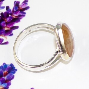 K&ouml;nnte beinhalten: Ein Sterling Silber Ring mit einem gro&szlig;en, facettierten, bernsteinfarbenen Edelstein. Der Ring hat ein schlichtes Band und eine Zargenfassung. Der ovale Edelstein f&auml;ngt das Licht ein und zeigt seine warmen Farbt&ouml;ne. Der Ring ist vor einem wei&szlig;en Hintergrund platziert.