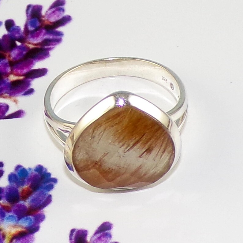 K&ouml;nnte beinhalten: Ein silberner Ring mit einem tropfenf&ouml;rmigen, hellbraunen Edelstein. Der Edelstein hat ein strukturiertes, faseriges Aussehen. Der Ring ist vor einem wei&szlig;en Hintergrund mit lila Blumen in der Ecke platziert. Der Ring ist ein Schmuckst&uuml;ck.