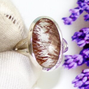 May include: A silver ring featuring an oval-shaped gemstone with a clear base and reddish-brown inclusions. The ring has a simple band and a bezel setting that holds the stone securely. The background is a soft white fabric and purple flowers.