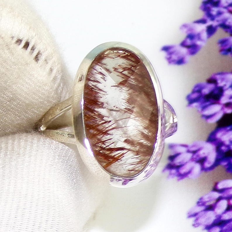 May include: A silver ring featuring an oval-shaped gemstone with a clear base and reddish-brown inclusions. The ring has a simple band and a bezel setting that holds the stone securely. The background is a soft white fabric and purple flowers.