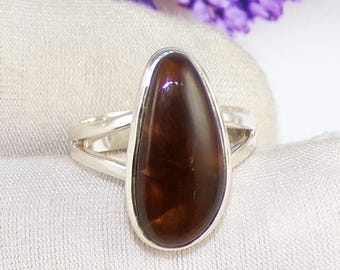 May include: A sterling silver ring featuring a large, teardrop-shaped, polished brown gemstone. The gemstone is set in a bezel setting and has a split shank design. The ring is displayed on a white surface with purple flowers in the background.