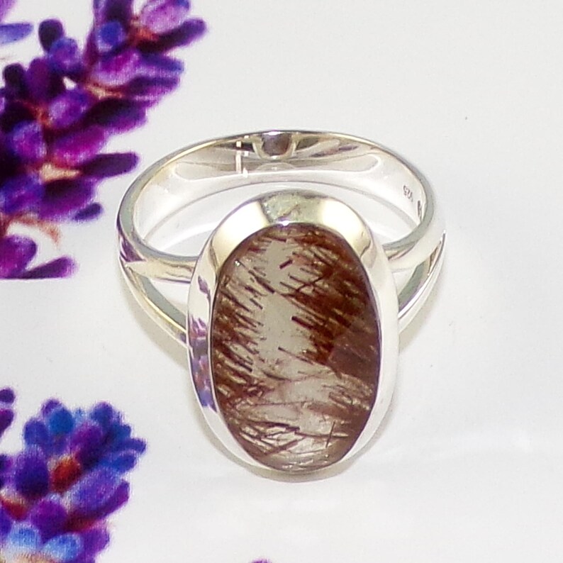 May include: A silver ring with an oval-shaped gemstone. The gemstone is a translucent light brown with dark brown inclusions. The ring is set against a white background with purple flowers in the corner.