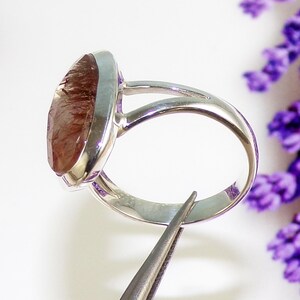 May include: A silver ring with a large, oval-shaped, reddish-brown gemstone. The ring's band splits into two at the top, creating a unique design. The gemstone is faceted, catching the light and displaying a variety of colors. The ring is held by tweezers.