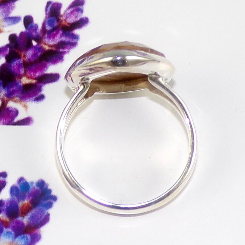 K&ouml;nnte beinhalten: Ein Silberring mit einem gro&szlig;en, facettierten, oval geformten Edelstein. Der Edelstein hat eine hellbraune Farbe. Der Ring hat ein schlichtes, poliertes Silberband. Der Ring ist vor einem wei&szlig;en Hintergrund mit lila floralen Akzenten platziert.