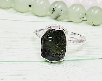 Peut inclure: Une bague en argent avec une pierre précieuse vert foncé taillée à la main. La bague est posée sur une surface en bois blanc.