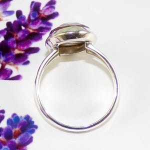 May include: A silver ring with a large, faceted, oval-shaped gemstone. The gemstone has a pale green hue. The ring is set on a white surface, with a blurred purple floral element in the background.