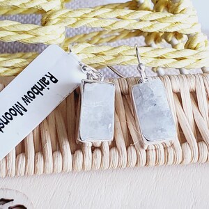Puede incluir: Un par de pendientes de plata con piedras rectangulares de piedra lunar blanca. Los pendientes están sobre una superficie de mimbre tejido con una etiqueta blanca que dice "Rainbow Moon".