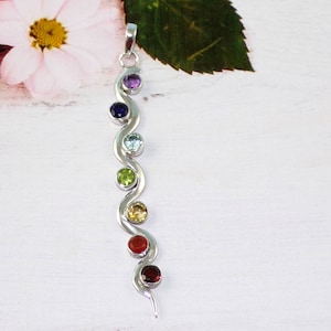 May include: A silver pendant featuring seven gemstones in a rainbow colour sequence. The pendant is shaped like a wavy line.
