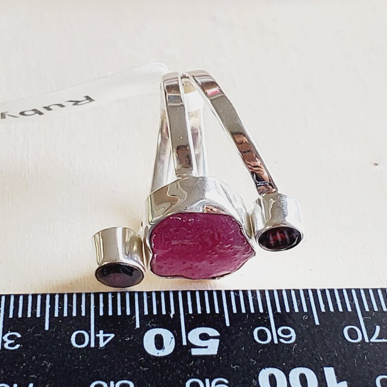 Peut inclure: Une bague en argent avec une grosse pierre de rubis rose brute et deux petites pierres rouge fonc&eacute;. La bague est pos&eacute;e sur une r&egrave;gle noire avec des graduations en pouces.