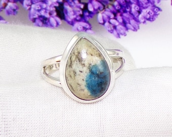 Peut inclure: Bague en argent ornée d'une pierre en forme de goutte d'eau, mélange de beige, blanc et bleu. La bague présente un design à tige fendue. Fond blanc avec des fleurs violettes.