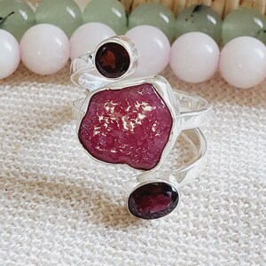 May include: A silver ring featuring a large, irregularly shaped, pink ruby gemstone and two smaller, round, dark red garnet gemstones.