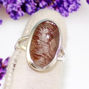 May include: A silver ring featuring an oval-shaped gemstone with reddish-brown inclusions. The gemstone is set in a polished silver bezel and has a faceted surface. The ring is displayed against a white background with purple flowers.