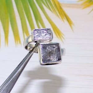May include: A silver ring with two square-shaped meteorite inlays. The inlays have a unique, textured surface.