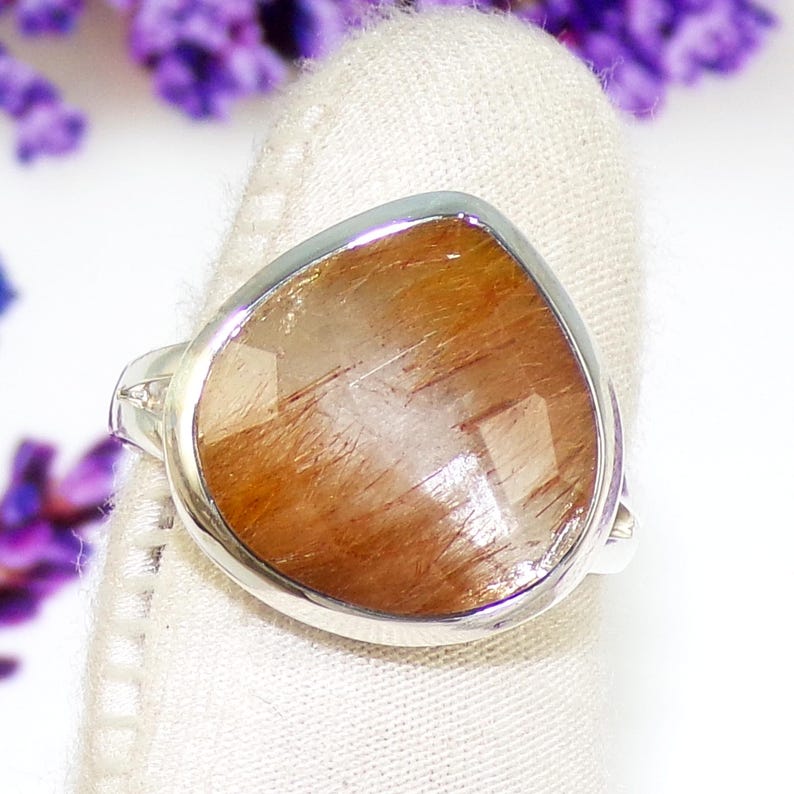 K&ouml;nnte beinhalten: Ein Silberring mit einem gro&szlig;en, tropfenf&ouml;rmigen Edelstein in warmem, rotbraun mit inneren Streifen. Der facettierte Stein f&auml;ngt das Licht ein. Der Ring liegt auf einem wei&szlig;en Hintergrund mit lila Blumen.