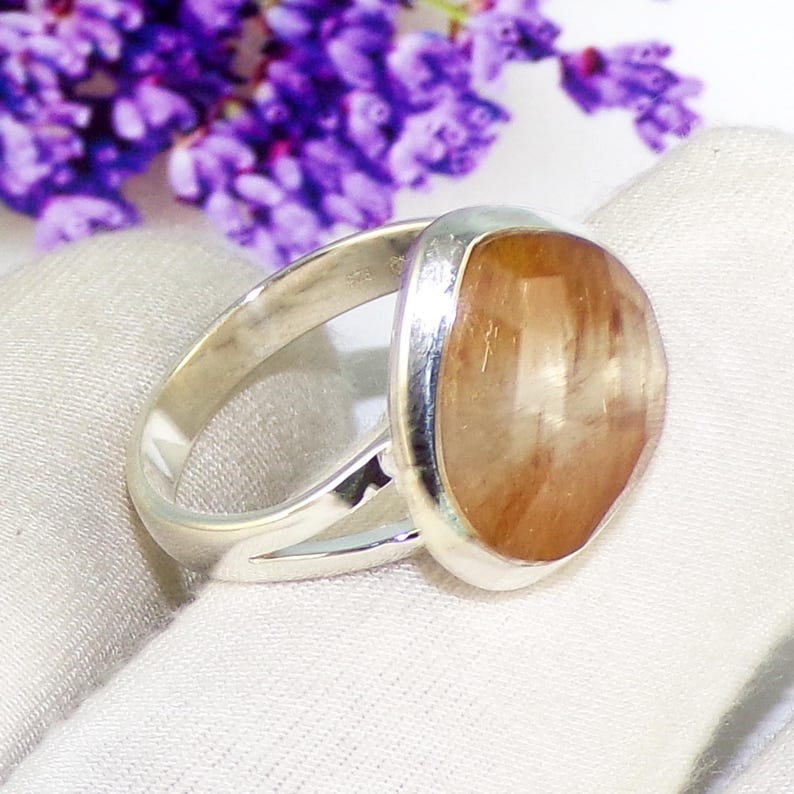 K&ouml;nnte beinhalten: Silberring mit einem gro&szlig;en, ovalen, hellbraunen Edelstein. Der Ring hat ein geteiltes Schaftdesign. Der Hintergrund ist wei&szlig; mit lila Blumen.
