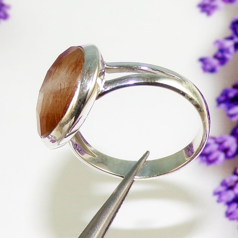 K&ouml;nnte beinhalten: Ein Silberring mit einem gro&szlig;en, oval geformten, facettierten Edelstein. Der Edelstein hat braune und orange Farbt&ouml;ne. Der Ring ist vor einem wei&szlig;en Hintergrund mit lila Blumen platziert.