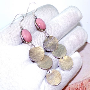 May include: A pair of silver dangle earrings with pink oval stones and multiple silver discs. The earrings are hanging from a white surface.