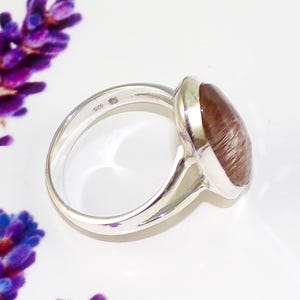 May include: A sterling silver ring with a large, oval-shaped, reddish-brown gemstone. The ring has a polished finish and a simple band. The gemstone has a unique, textured appearance. The ring is set against a white background with purple flowers.