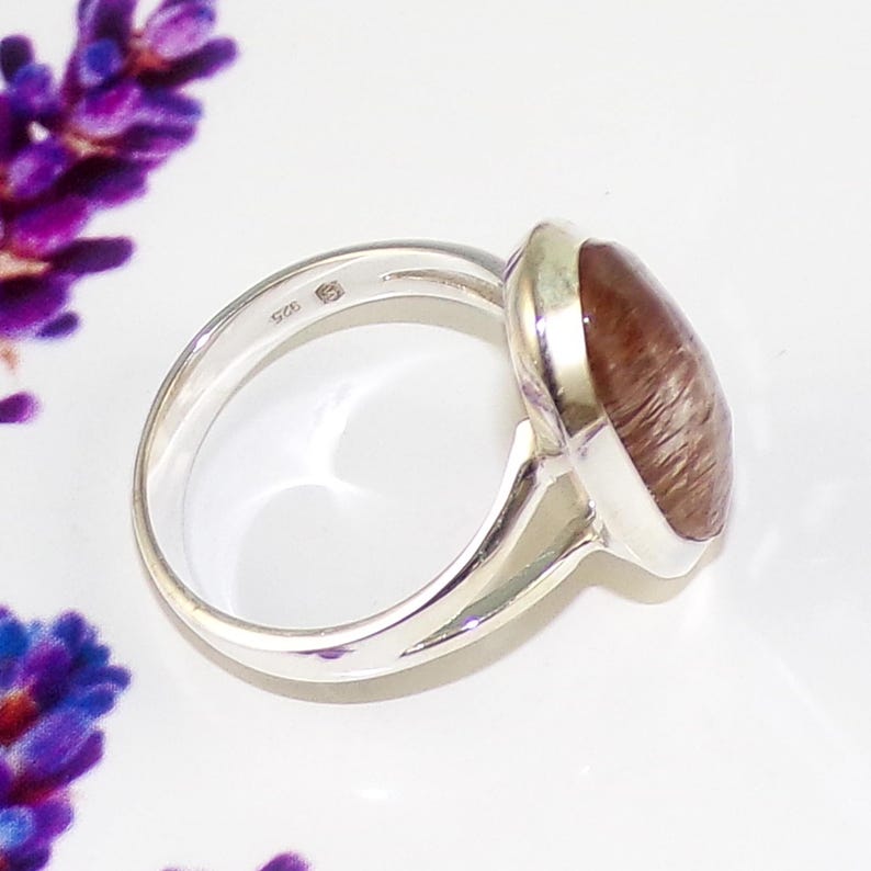 May include: A sterling silver ring with a large, oval-shaped, reddish-brown gemstone. The ring has a polished finish and a simple band. The gemstone has a unique, textured appearance. The ring is set against a white background with purple flowers.