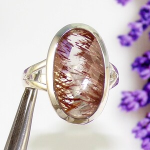 May include: A silver ring featuring an oval-shaped gemstone with a clear base and intricate reddish-brown inclusions. The ring has a polished silver band and a bezel setting that highlights the stone's unique pattern.