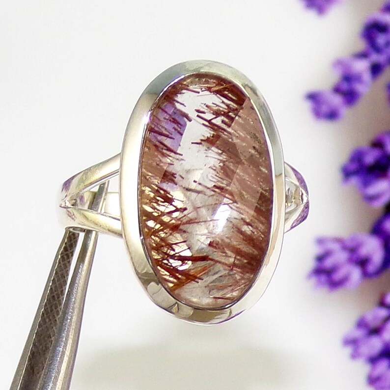 May include: A silver ring featuring an oval-shaped gemstone with a clear base and intricate reddish-brown inclusions. The ring has a polished silver band and a bezel setting that highlights the stone's unique pattern.