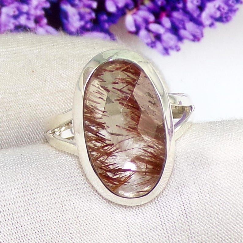 May include: A silver ring featuring an oval-cut gemstone with reddish-brown inclusions. The ring has a polished silver band and a bezel setting. The background includes a soft white fabric and blurred purple flowers.
