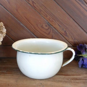 May include: White enamel chamber pot with a green rim and a handle. The pot is sitting on a wooden surface.