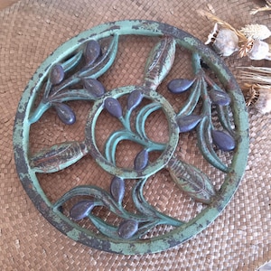 May include: A round, distressed metal olive branch design in shades of teal, brown, and purple. The decorative piece is placed on a woven mat, with dried plants in the background. The metal piece has a rustic, vintage aesthetic.