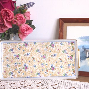Può includere: Vassoio rettangolare in ceramica con motivo floreale rosa, blu e giallo. Il vassoio ha manici dorati e un bordo blu. Lo sfondo include un vaso di rose rosa e un acquerello incorniciato.