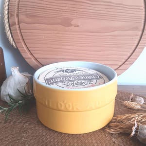 Peut inclure: Plat à fromage en céramique jaune avec un couvercle. Le couvercle porte une étiquette blanche avec le texte "Creme de l'Argent" en lettres noires. Le plat est sur une surface tissée.