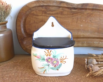 Puede incluir: Un salero de cerámica blanca con tapa marrón oscuro y ribete azul. El salero presenta un diseño floral en rosa, verde y naranja. El salero está colgado en una pared.