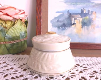 May include: A white ceramic jar with a gold trim and a lid. The jar has a floral design and is sitting on a white crocheted doily.