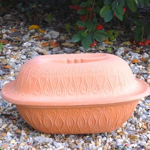 Puede incluir: Una olla de cerámica de color terracota con tapa. La tapa tiene las palabras "Schlemmertopf" en relieve. La olla tiene un patrón decorativo de llamas en los lados.