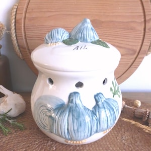 Peut inclure: Pot à ail en céramique blanche avec des motifs d'ampoules d'ail bleues et le mot "AIL" sur le couvercle. Le pot a des trous d'aération et est décoré de feuilles vertes. Un article de cuisine décoratif.