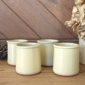 Puede incluir: Cuatro vasos de cerámica color crema con una forma ligeramente acampanada. Los vasos tienen un acabado liso y mate y una base de color marrón rojizo. Están dispuestos sobre una superficie de madera, lo que sugiere una estética rústica o campestre.