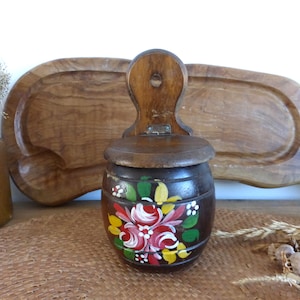 Puede incluir: Un salero o pimentero de madera para colgar en la pared con una tapa con bisagras. El salero está pintado con un diseño floral en tonos rojos, amarillos, verdes y blancos.