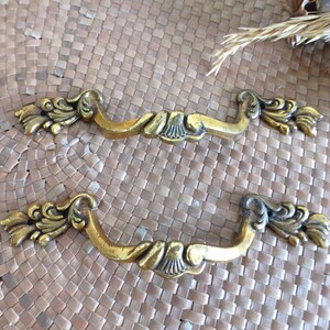 Puede incluir: Dos tiradores de cajón de color dorado ornamentados con un diseño de desplazamiento decorativo. Los tiradores tienen un diseño floral en cada extremo.