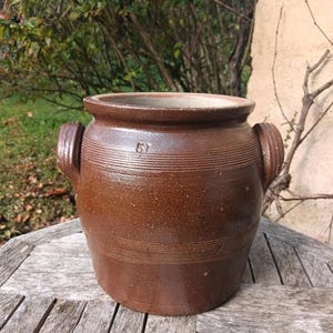 May include: A brown stoneware crock with two handles and a number "67" inscribed on the front. The crock is sitting on a wooden table.