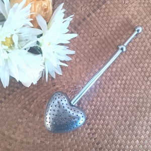 May include: A heart-shaped, silver tea infuser with a long handle and small perforations. The infuser is placed on a woven brown surface, with white flowers in the background. The tea infuser is designed for steeping loose leaf tea.