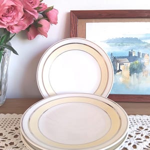 Puede incluir: Un juego de platos de cerámica blanca con un borde amarillo pálido y dorado. Los platos están apilados sobre una mesa con un tapete de encaje. Un jarrón de rosas rosas y una acuarela enmarcada están en el fondo.