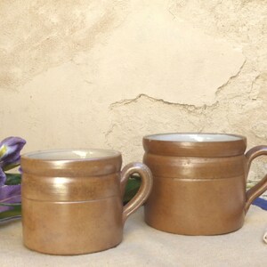 May include: Two brown ceramic mugs with handles. The mugs have a light brown exterior with a darker brown band around the middle. The interior of the mugs is white. The mugs are sitting on a white and blue striped surface.